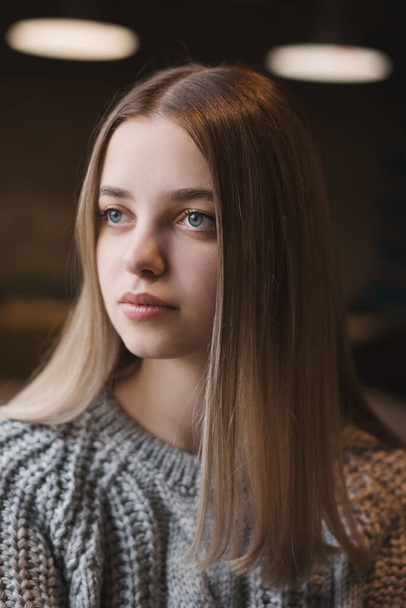 Πορτρέτο ενός έφηβου κοριτσιού με ξανθά μαλλιά και μπλε μάτια κάθεται σε μοντέρνο πλεκτό ζακέτα από γκρι χρώμα - Φωτογραφία, εικόνα