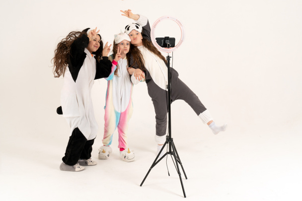 Chicas adolescentes lindas en kigurumi y máscaras para dormir sonriendo y dispara un video. Selfies. El teléfono está montado en un trípode y la lámpara del anillo brilla. - Foto, imagen