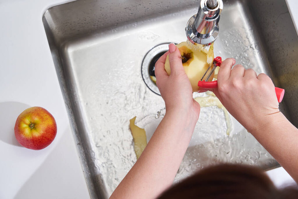 Καλλιεργημένη άποψη των γυναικείων χεριών ξεφλούδισμα μήλου πάνω από τη μηχανή διάθεσης απορριμμάτων τροφίμων - Φωτογραφία, εικόνα