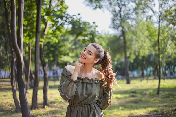 Portret szczęśliwej młodej dziewczyny w zielonej sukience, z pięknymi włosami. Spacer w letnim zielonym parku w słoneczny dzień. - Zdjęcie, obraz