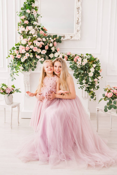 Blonde Mutter mit einer niedlichen Tochter in eleganten flauschigen Kleidern vor dem Hintergrund eines wunderschönen Interieurs mit Blumen. Das Bild der Prinzessinnen. Das Konzept der Feier zum Geburtstag und Frauentag - Foto, Bild