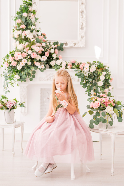 Beautiful blonde girl with long hair in a soft purple dress on a background of flowers posing. Cute baby model in the image of a Princess. She's wearing a full lilac dress. - Valokuva, kuva