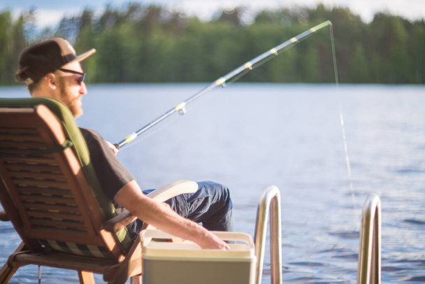 Hombre maduro pescando desde el muelle de madera cerca de casa de campo en el lago en Finlandia en verano
 - Foto, Imagen