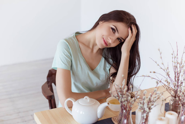 A beautiful brunette woman sits at a table with a mug of green tea and smoothes her hair smiling, flirting. Life style. - Photo, image