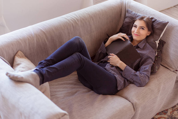 Beautiful brunette woman sitting on sofa with book, in jeans and shirt. Sexy adult woman of 30 years. Life style. - Photo, image