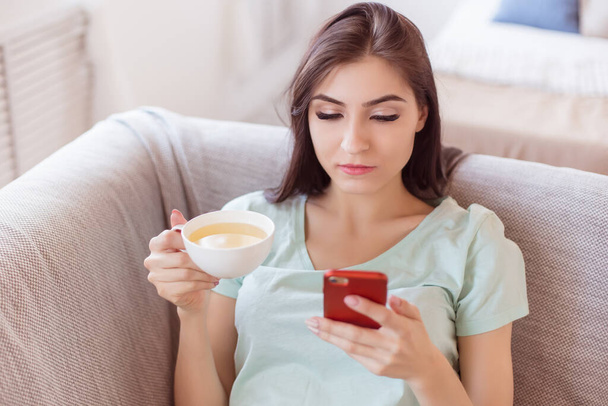 Beautiful brunette woman lying on the couch with a smartphone in her hands and a cup of green tea. Sexy adult woman of 30 years. Life style. - Foto, Bild