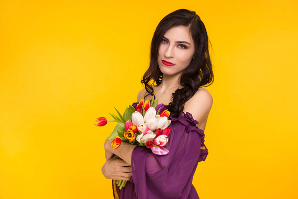 Beautiful brunette woman holding a bouquet of tulips in her hands on a yellow background. Female dressed in a purple, thin dress. March 8, the day matri. - Photo, Image