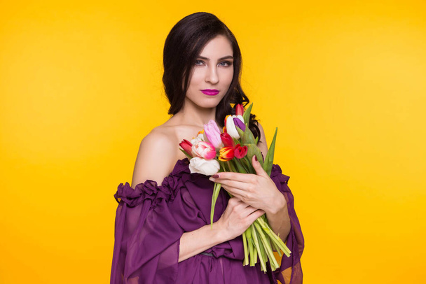 Beautiful brunette woman holding a bouquet of tulips in her hands on a yellow background. Female dressed in a purple, thin dress. March 8, the day matri. - Photo, image