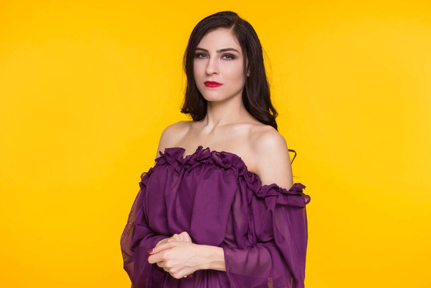 Beautiful brunette woman in a purple dress and bare shoulders on a yellow background. Copy space. - Φωτογραφία, εικόνα