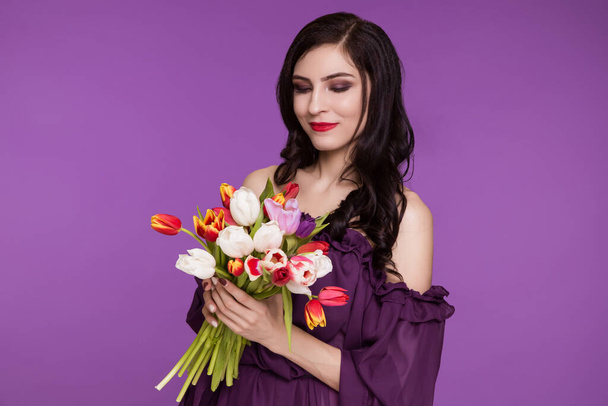 Beautiful young woman, brunette, stands on a purple background, in an air dress and a bouquet of tulips. International Women's Day, March 8. - Zdjęcie, obraz