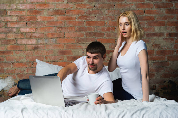 Young couple of guy and girl in bed and watching shocked news on laptop. - Фото, изображение