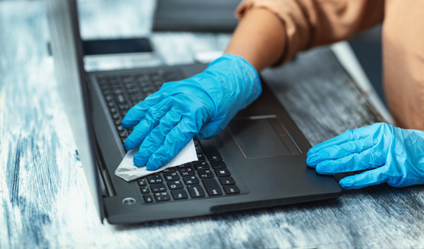 Karantinada mavi eldiven giyen bir kadın dizüstü bilgisayar temizliyor. - Fotoğraf, Görsel