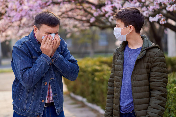 отец и сын в маске находятся в городе на открытом воздухе, цветущие деревья, весенний сезон, время цветения - концепция аллергии и охраны здоровья от пыльного воздуха
 - Фото, изображение