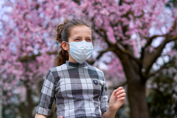 κορίτσι με μάσκα προσώπου είναι στην πόλη υπαίθρια, ανθίζοντας δέντρα, την άνοιξη, την εποχή της ανθοφορίας - έννοια των αλλεργιών και της προστασίας της υγείας από σκονισμένο αέρα - Φωτογραφία, εικόνα