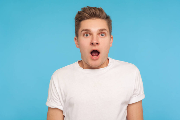 Wow, unbelievable! Portrait of astonished man in casual white t-shirt looking at camera with mouth open in amazement, scared and shocked by crazy news. indoor studio shot isolated on blue background - Photo, Image