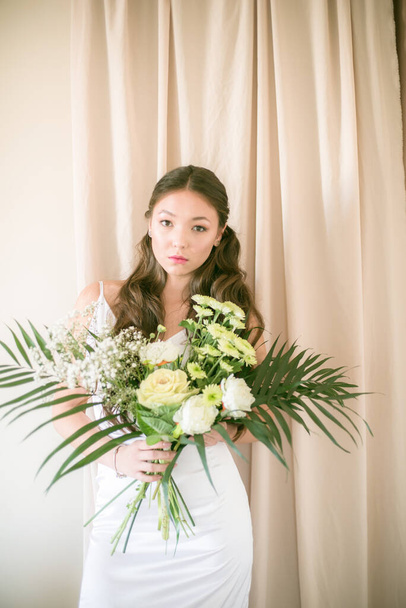 Cute tender young bride of Asian appearance with long hair in a white dress with a wedding bouquet of flowers in the studio - Fotó, kép