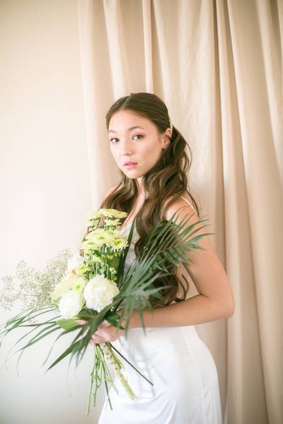Cute tender young bride of Asian appearance with long hair in a white dress with a wedding bouquet of flowers in the studio - Фото, зображення