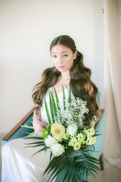 Cute tender young bride of Asian appearance with long hair in a white dress with a wedding bouquet of flowers in the studio - Foto, imagen