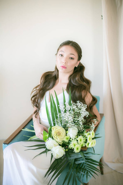 Ніжна молода наречена азіатського вигляду з довгим волоссям у білій сукні з букетом квітів у студії - Фото, зображення