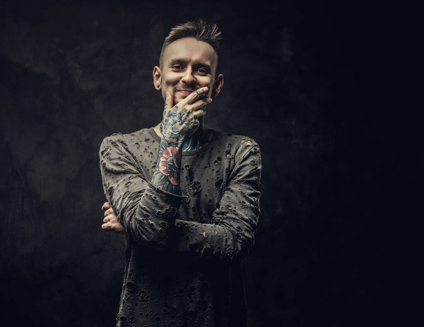 Inked young adult male wearing ripped jumper posing in a dark studio on a grey background - Photo, Image