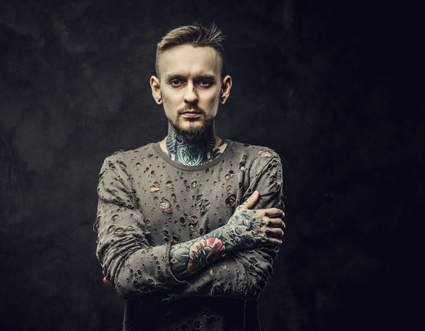 Ανδρικό μοντέλο με τατουάζ που ποζάρει σε σκούρο γκρι φόντο φορώντας σκισμένο μακρυμάνικο πουλόβερ - Φωτογραφία, εικόνα