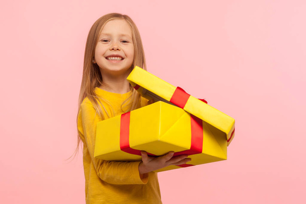 Child satisfied with good present. Portrait of charming funny little girl opening gift box and smiling to camera, celebrating birthday, Christmas holiday. studio shot isolated on pink background - Photo, Image