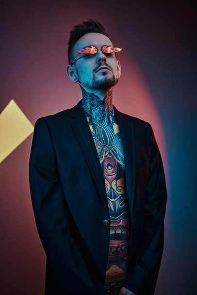 Tetováló művész férfi modell pózol egy neon stúdióban félmeztelen tetovált testtel, szmokinggal és napszemüveggel. Függőleges portré - Fotó, kép