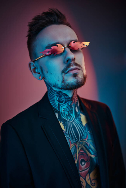 Крутой мужчина-татуировщик позирует в неоновой студии с полуобнаженным телом, смокингом и солнечными очками. Вертикальный портрет
 - Фото, изображение