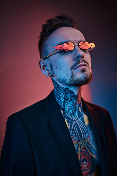 Modèle masculin tatoueur posant dans un studio néon avec corps tatoué à moitié nu, smoking et lunettes de soleil. Portrait vertical gros plan
 - Photo, image