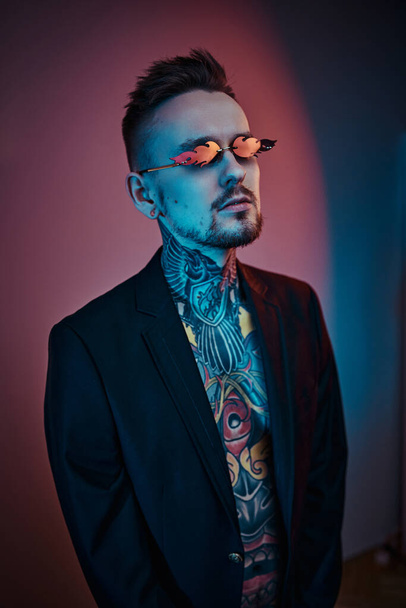 Modelo masculino de aparência alternativa posando em um estúdio de néon com corpo tatuado semi-nu, smoking e óculos de sol. Retrato vertical
 - Foto, Imagem