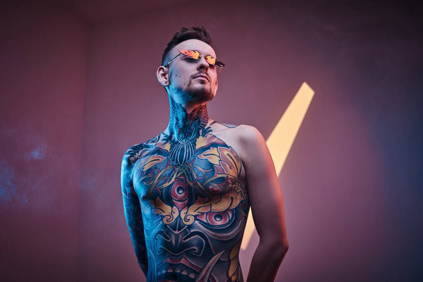 Mustettu miesmalli poseeraa neonstudiossa puolialastomalla tatuoidulla vartalolla ja aurinkolaseilla. Pystysuora muotokuva
 - Valokuva, kuva