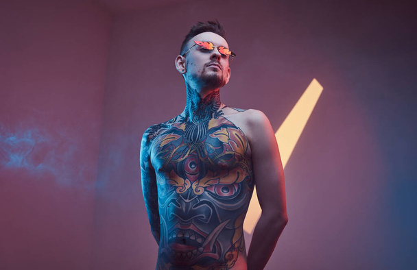 Inkoustovaný mužský model pózující v neonovém studiu s polonahým tetovaným tělem a slunečními brýlemi. Svislý portrét - Fotografie, Obrázek