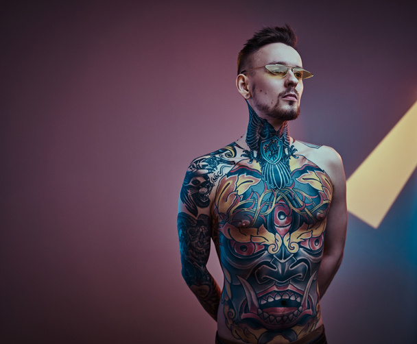 Мужская модель-татуировщик позирует в неоновой студии с полуобнаженным телом и солнечными очками. Вертикальный портрет
 - Фото, изображение