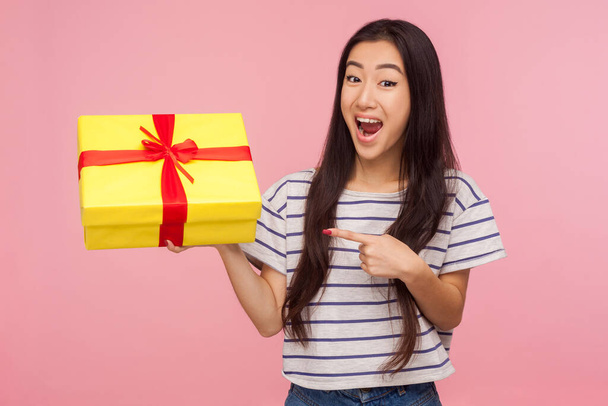 Awesome birthday present! Amazed satisfied pretty girl with brunette hair in striped t-shirt pointing at gift box and looking with surprised expression. indoor studio shot isolated on pink background - Φωτογραφία, εικόνα