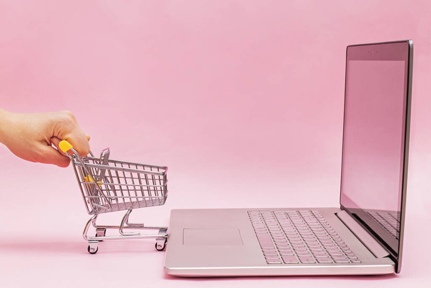 Vrouwen hand houden mini winkelwagentje in de buurt van de geopende laptop op roze achtergrond. - Foto, afbeelding
