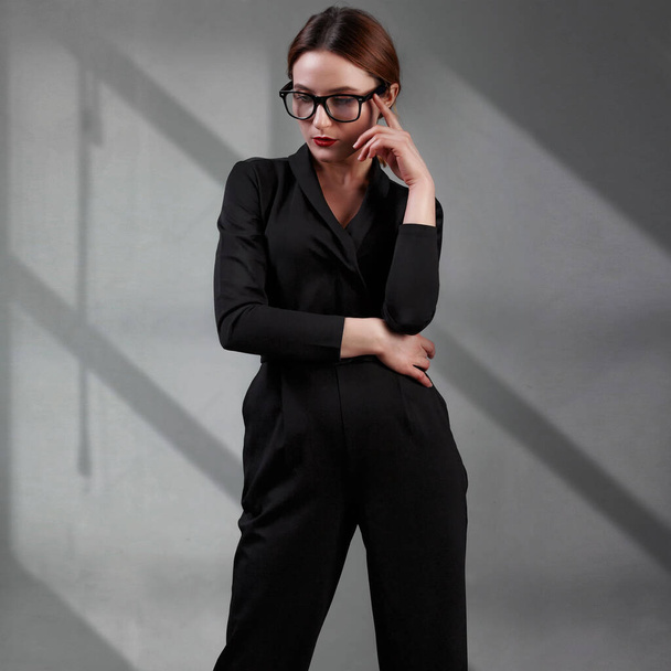 elegant business woman in black classic overalls and glasess posing over gray background. Happy satisfied relaxed elegant woman enjoy her work - Foto, Imagen