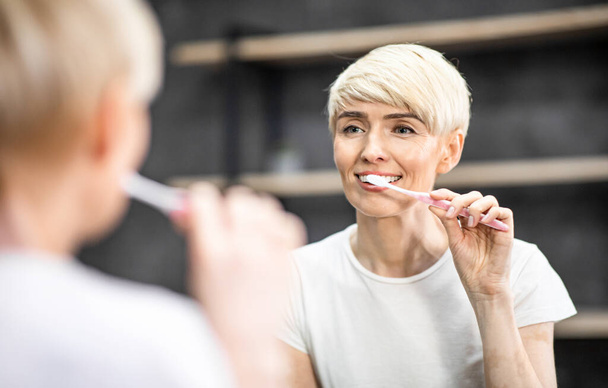 Nainen harjaamalla hampaat hymyilee heijastus peilissä kylpyhuoneessa
 - Valokuva, kuva