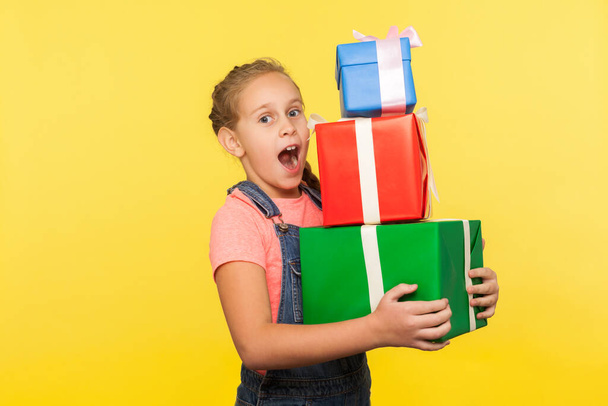 Portrait of surprised little girl with braid in denim overalls holding lot of gift boxes and looking with amazement, shocked by many presents on her birthday. studio shot isolated on yellow background - Фото, изображение