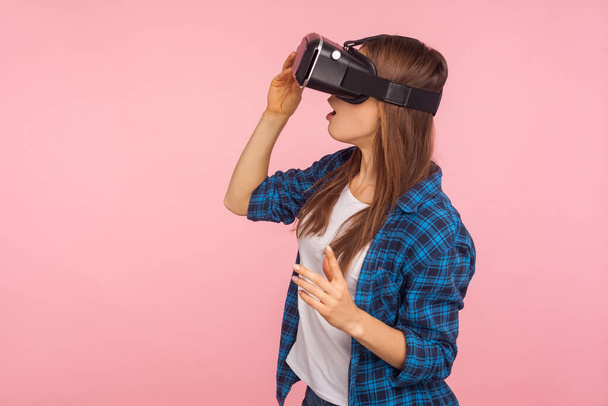 Side view of gamer girl in checkered shirt wearing vr headset, playing virtual reality game with amazed facial expression, surprised by innovative technology. studio shot isolated on pink background - Φωτογραφία, εικόνα