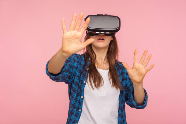 Portrait of young gamer woman in checkered shirt wearing vr headset and touching air with hands, playing virtual reality game with serious concentrated expression. indoor studio shot, isolated - Foto, afbeelding