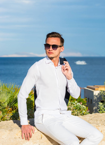 Junger gutaussehender Mann in Anzug und weißer Hose vor Meeresgrund und Palmen. Modisches Modell - Foto, Bild