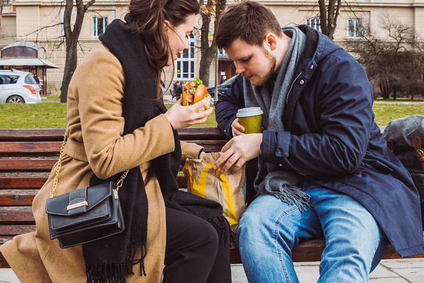 νεαρό ενήλικο ζευγάρι που τρώει φαστ φουντ στον πάγκο του πάρκου της πόλης. έννοια του τρόπου ζωής - Φωτογραφία, εικόνα