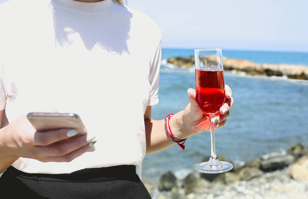 Relaxed young female in casual wear holding glass of rose champagne and holding mobile phone enjoying fresh breeze while standing aboard of ship during sea trip - Photo, image