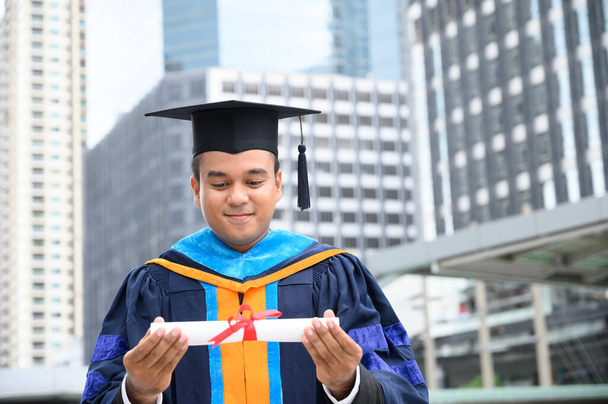 Buon diploma. Happy Asain uomo in abiti di laurea in possesso di diploma in mano su sfondo urbano della città
. - Foto, immagini