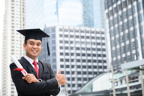 Glücklicher Absolvent. Glücklicher Asain Mann im Abschlusskleid mit Diplom in der Hand vor städtischem Hintergrund. - Foto, Bild