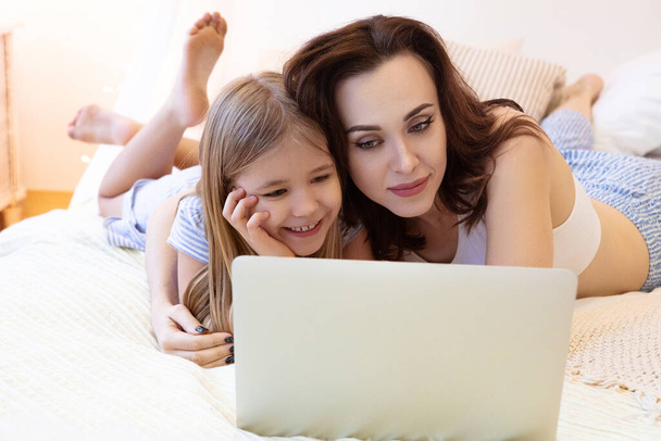 Mujer adulta y chica feliz tumbada en la cama y viendo películas en el portátil mientras pasan el día de fin de semana en casa juntos
 - Foto, Imagen