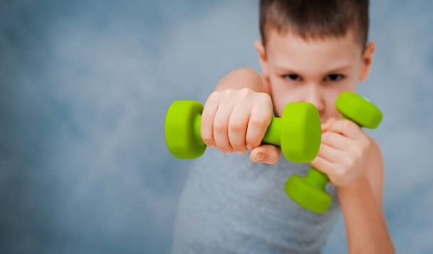 Cry sport bébé garçon montre émotionnellement son bras biceps force musculaire avec haltères sur un fond bleu gris - Photo, image