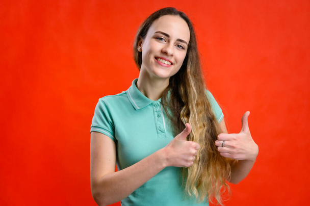 Portrait of a young beautiful girl on a red background. A model with long hair is talking to the camera, showing hands. - Foto, imagen