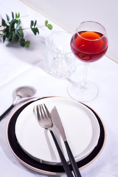 Посуда и украшения для сервировки праздничного стола. Тарелки, бокал красного вина и столовые приборы с зелеными листьями на белом текстильном фоне
. - Фото, изображение