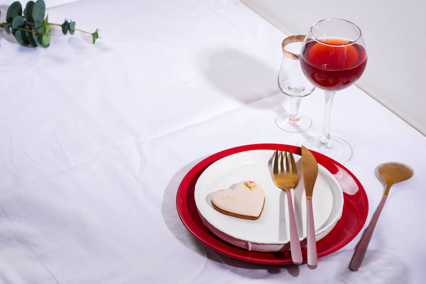 Tableware and decorations for serving a festive table. Plates, red wine glass and cutlery with heart cookie on white textile background. - Photo, Image